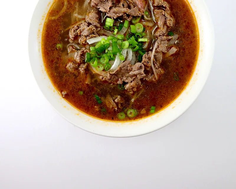 Bowl of sate beef noodle soup in a red-orange spicy broth with sliced beef and green onion